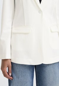 Blazer blanc ajusté avec un seul bouton, revers crantés et deux poches avant, associé à un jean en denim bleu. Texture lisse et coupe structurée.