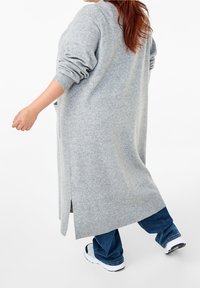 Lång grå cardigan i mjukt stickad tyg, med rullade ärmar, sidofickor och en avslappnad design, kombinerad med jeansbyxor och vita sneakers.