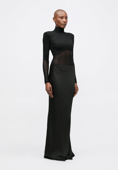 Bluza neagră cu mâneci lungi, cu panouri transparente la nivelul trunchiului, combinată cu o fustă neagră ajustată, până la podea, cu textură fină, design minimalist.