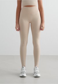 Leggings beige a vita alta realizzati in tessuto liscio e elastico, abbinati a scarpe da ginnastica bianche con dettagli grigi e una suola testurizzata.
