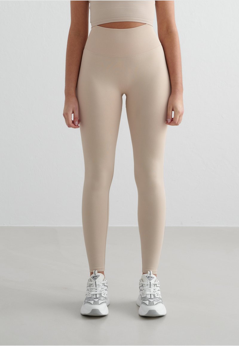 Leggings beige a vita alta realizzati in tessuto liscio e elastico, abbinati a scarpe da ginnastica bianche con dettagli grigi e una suola testurizzata.