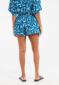 Shorts bleus avec des motifs abstraits blancs, taille élastique et coupe décontractée. Associés à des tongs noires simples.
