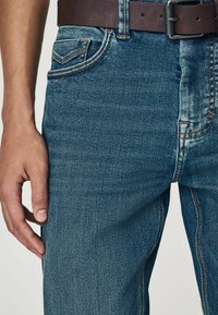 Blugi din denim albastru cu o textura estompată, având o curea din piele maro și cusături contrastante de-a lungul buzunarelor.