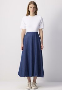 Un haut blanc à manches courtes associé à une jupe maxi en denim bleu évasée, présentant une texture lisse et un subtil motif diagonal, complété par des baskets décontractées.