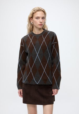 Femme blonde portant un pull argyle foncé avec des lignes bleues et une jupe marron foncé, debout devant un fond clair uni.