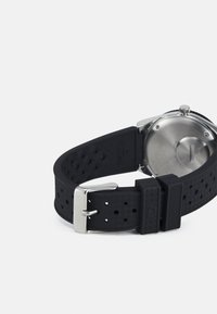 Black silicone watch strap with circular stainless steel case. Features perforated texture and silver metal buckle with embossed branding.