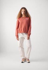 Sweatshirt couleur rouille à col rond, avec un petit logo sur la poitrine et un emblème Jordan sur la manche. Associé à un pantalon cargo blanc et des baskets.