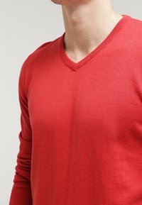 Pull rouge en V fabriqué en tissu fin, avec des manches longues et des bords côtelés autour de l'encolure et des poignets. Texture lisse.