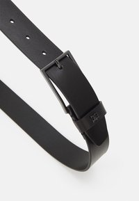 Cintura nera in pelle con una superficie liscia, dotata di una fibbia metallica rettangolare e multiple perforazioni per la regolazione della misura.