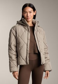 Giacca puffer trapuntata beige con colletto alto e cappuccio, dotata di zip frontale e bottoni a pressione, abbinata a un top in maglia marrone e leggings.