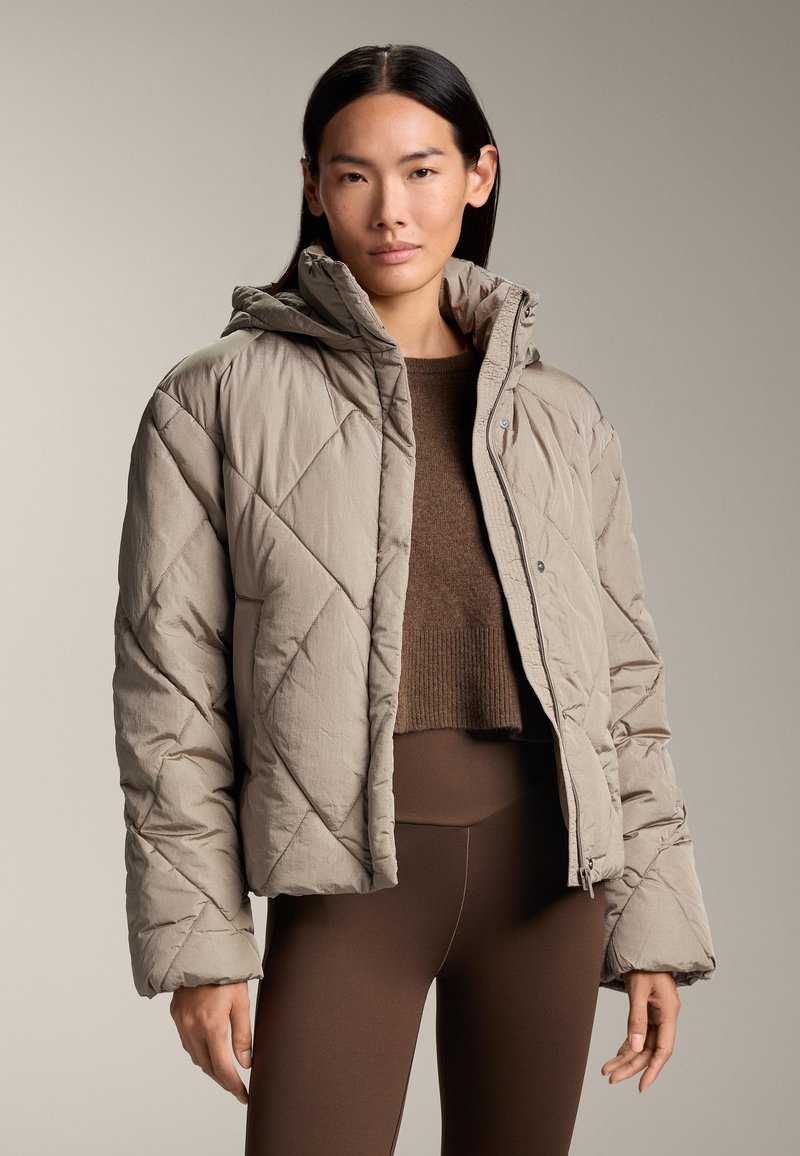 Giacca puffer trapuntata beige con colletto alto e cappuccio, dotata di zip frontale e bottoni a pressione, abbinata a un top in maglia marrone e leggings.