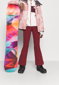 Ein Snowboard mit einem mehrfarbigen Design lehnt an einer Person in einer rosa Batikjacke, weißem Fleece und kastanienbraunen Schneehosen und Stiefeln.