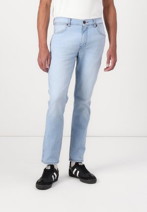 Homme portant un jean slim bleu clair, des chaussettes blanches et des baskets noires avec des rayures blanches, debout devant un fond blanc uni.