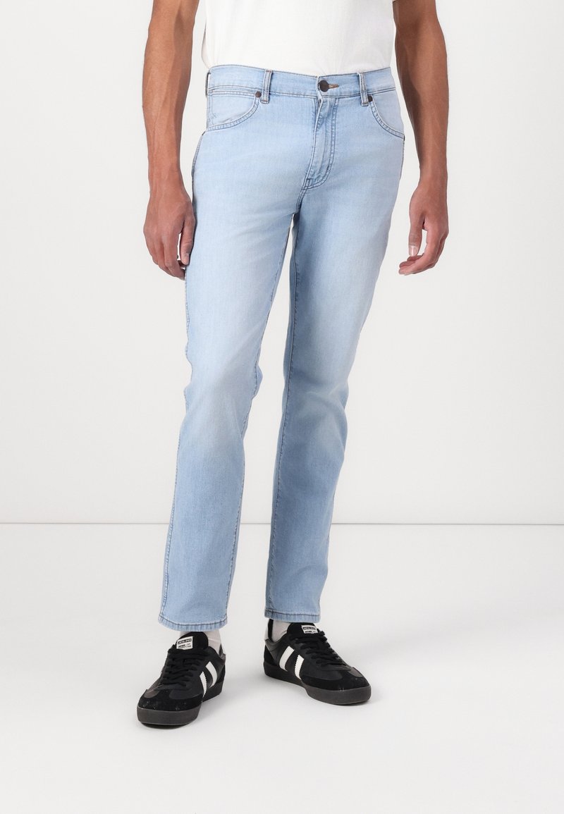 Homme portant un jean slim bleu clair, des chaussettes blanches et des baskets noires avec des rayures blanches, debout devant un fond blanc uni.