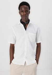 Camicia bianca di lino a maniche corte con taschino e colletto button-down, abbinata a un bandana nera attorno al collo. Texture liscia.