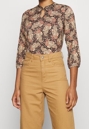 Kvinde iført en blomsterprikket paisley langærmet bluse med nøglehul ved halsen og højtaljede beige bukser.