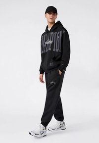 Sudadera con capucha negra con un gran logotipo gris de "Champion", combinada con pantalones jogger negros que llevan el texto "Champion" bordado y zapatillas deportivas blancas.
