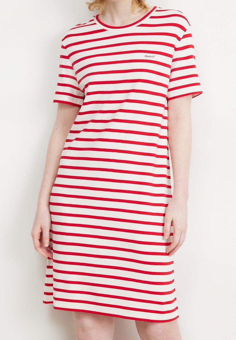 Femme portant une robe blanche mi-longue avec des rayures horizontales rouges et des manches courtes, se tenant devant un fond clair.