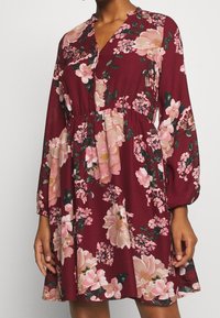 Vestido floral marrón con mangas largas, escote en V, cintura elástica y patrones de flores rosas. Tejido suave con un diseño ligeramente fluido.