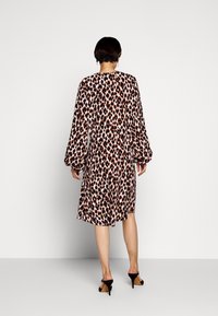 Kobieta w sukience w panterkę o długości do kolan z długim rękawem oraz czarnych sandałach na wysokim obcasie, stojąca pod gładką białą ścianą.