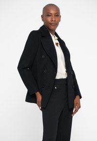 Διπλής όψης μαύρο παλτό από μίγμα μαλλιού με μεγάλους μπροστινούς κουμπιά και μυτερό γιακά, φορεμένο πάνω από λευκό πουκάμισο και μαύρο παντελόνι. Καθαρός σχεδιασμός.