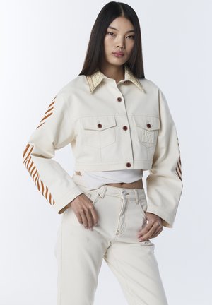 Joven mujer con chaqueta corta color crema con rayas marrones en las mangas y pantalones de talle alto a juego, posando frente a un fondo blanco.