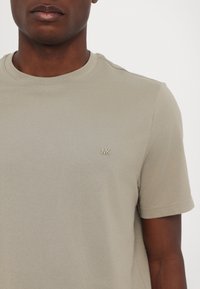 Kortermet t-skjorte i lys beige bomull med rund hals, som har en liten beige "MK"-logo på venstre bryst. Glatt tekstur.