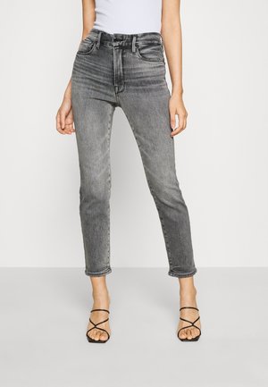 Vrouw draagt hooggetailleerde vervaagde zwarte skinny jeans en zwarte sandalen met hak en bandjes, staand tegen een effen lichte achtergrond.