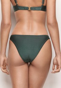 Bas de bikini côtelé vert foncé avec une taille basse. Il présente une texture lisse et une couverture minimale, conçu pour un look ajusté.