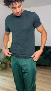 Ung mann med krøllete hår som justerer en mørk, tettsittende t-skjorte, iført løse grønne bukser, stående i en stue med planter og en mørk sofa.