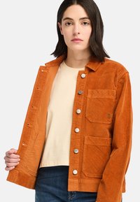 Orange kordjacka med knappstängning framtill, två bröstfickor och metallknappar, matchad med en krämfärgad skjorta och mörka jeans.