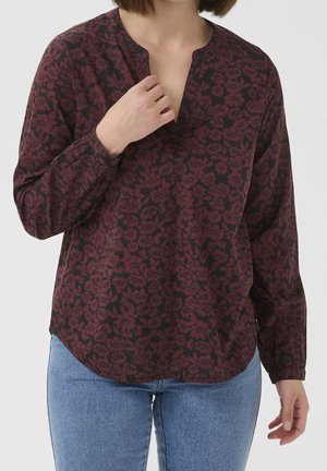 Kvinde iført en mørkerød bluse med lange ærmer og sorte blomsterprint samt lyseblå jeans, hånd rører ved udskæringen på blusen.