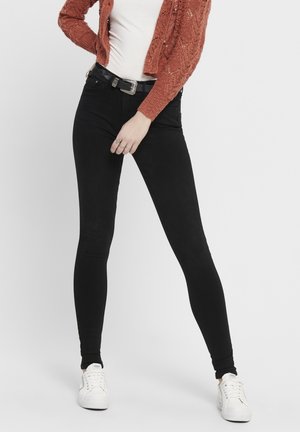 Femme portant un jean skinny noir, des baskets blanches, un haut blanc et un cardigan en maille couleur rouille, debout jambes écartées sur fond blanc.