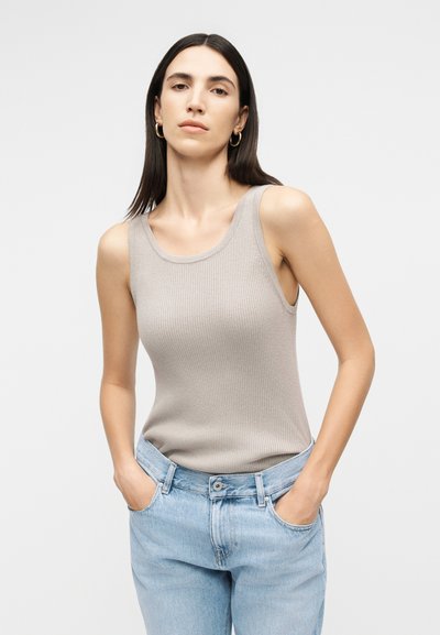 Γυναίκα με ίσια σκούρα μαλλιά που φοράει χρυσούς κρίκους στα αυτιά, μια μπεζ ριγωτή φανέλα χωρίς μανίκια και ανοιχτό μπλε τζιν, με τα χέρια στις τσέπες.