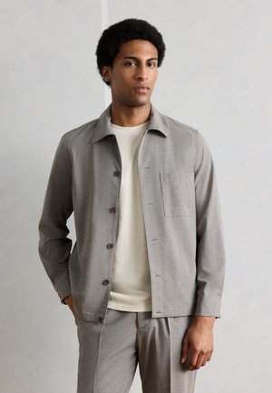 Άνδρας με κοντά σγουρά μαλλιά, φορώντας ανοιχτό γκρι πουκάμισο-σακάκι με κουμπιά πάνω από μια μπεζ μπλούζα και ταιριαστά γκρι παντελόνια.