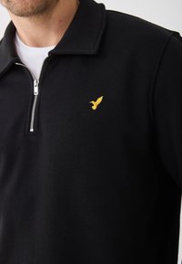 Felpa nera con colletto zip, dotata di un piccolo logo di un uccellino ricamato di colore giallo sul petto sinistro. Il tessuto sembra morbido e texturizzato.