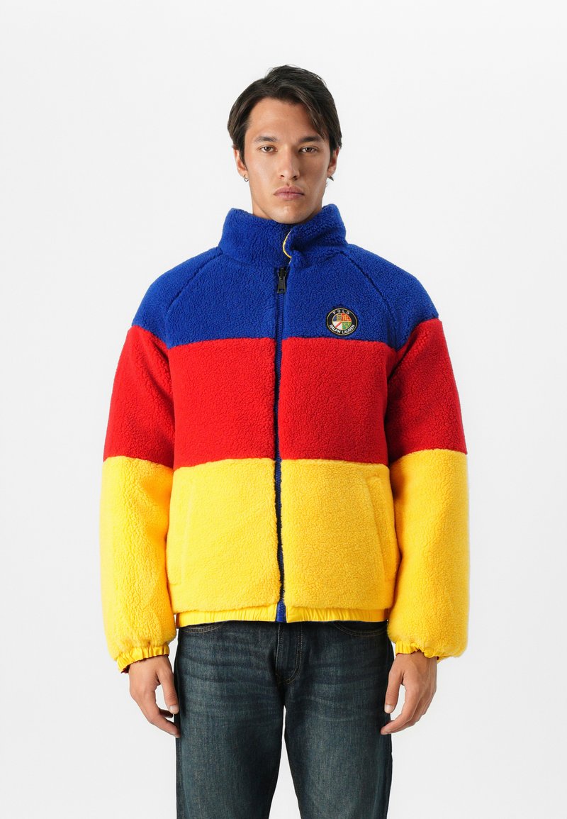 Mężczyzna w niebieskiej, czerwonej i żółtej polarowej kurtce w kolorowe bloki z wysokim kołnierzem i zamkiem, stojący na jednolitym białym tle.