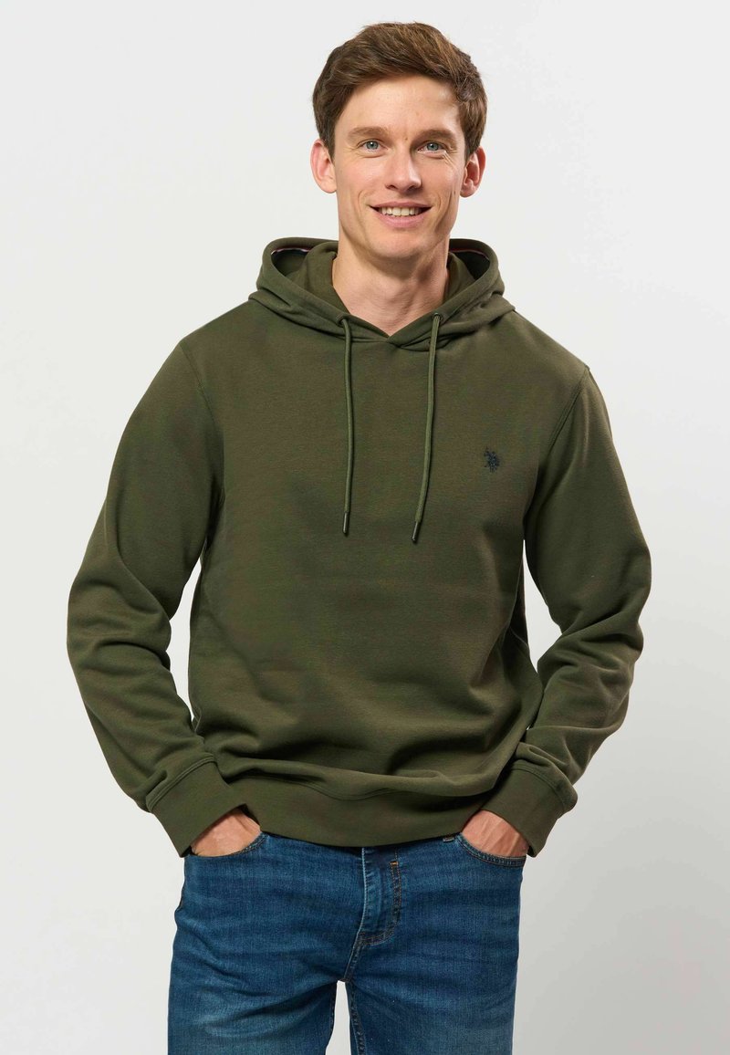 Jeune homme souriant, portant un sweat à capuche vert olive et un jean bleu, debout les mains dans les poches devant un fond clair uni.