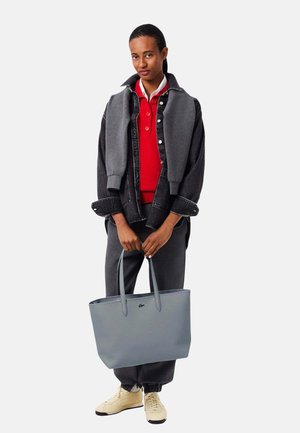 Personne portant un polo rouge et une veste noire, tenant un grand cabas gris, vêtue d’un pantalon foncé et de baskets beiges sur fond blanc.