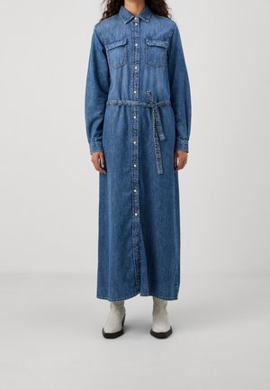 Kvinde iført en lang blå denim skjortekjole med knapper, brystlommer, et bundet bælte i taljen og hvide ankelstøvler på ensfarvet baggrund.