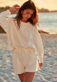 Hvid, oversized sweater med ribstrikkede manchetter og matchende shorts med snoretræk, lavet af blødt stof, båret mod en strandbaggrund ved solnedgang.