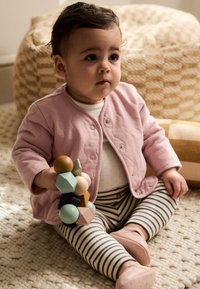 Lyserød quiltet cardigan, hvid skjorte med polkaprikker, stribede bukser. Holder et farverigt træstablelegetøj. Tekstureret tæppe og vævet puf i baggrunden.