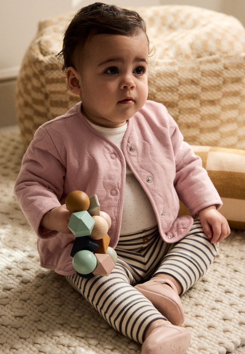 Cardigan matelassé rose, chemise blanche à pois, pantalon rayé. Tenant un jouet empilable en bois coloré. Tapis texturé et pouf tissé en arrière-plan.