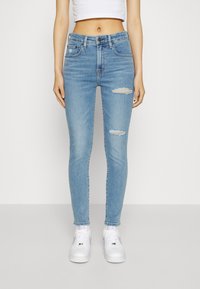 Vaqueros ajustados de color azul claro con detalles de rodillas rasgadas, de tiro medio, cinco bolsillos y una textura suave, combinados con zapatillas blancas.