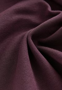 Tessuto bordeaux con una superficie liscia, che mostra morbidi drappeggi e sottili motivi a maglia. La luce si riflette delicatamente sulla superficie.