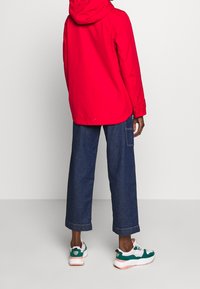 Chaqueta roja con capucha, vaqueros oscuros holgados y zapatillas multicolores. La tela es suave, con costuras visibles y un ajuste relajado.