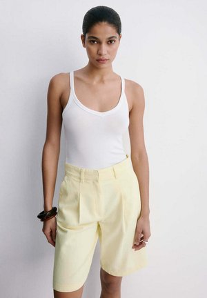 Mujer con cabello oscuro que lleva una camiseta sin mangas blanca y pantalones cortos hasta la rodilla de color amarillo pálido, de pie frente a un fondo blanco liso.