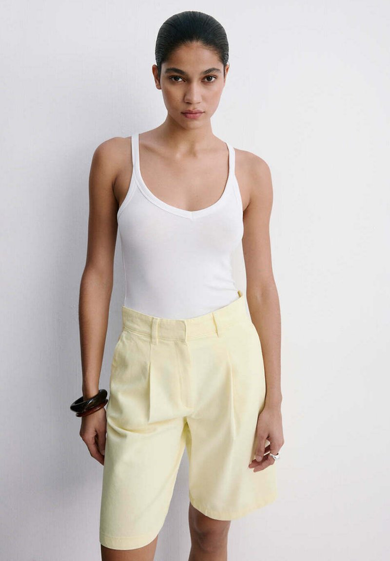 Femme aux cheveux foncés portant un débardeur blanc et un short jaune pâle jusqu'aux genoux, debout devant un fond blanc uni.