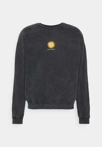 Svart långärmad crewneck sweatshirt med en liten gul solgrafik och texten "KAOTIKO SOCIETY" på bröstet.