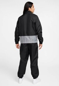 Μαύρο αδιάβροχο με γκρι ταινία στη μέση, ελαστικά μανίκια και ταιριαστή μαύρη παντελόνα με σούρες στους αστραγάλους. Συνδυάζεται με λευκά αθλητικά παπούτσια.